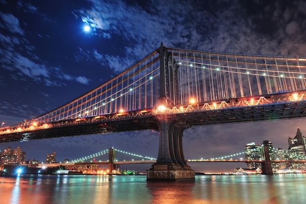 Brooklyn Bridge and Manhattan Bridge over East River at night with moon in New York City Manhattan with lights and reflections.