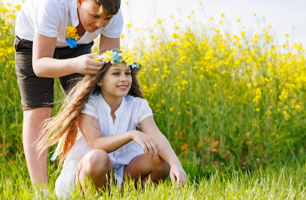 Brother braids ribbons in Ukrainian wreath with flowers on head of sister on meadow against backdrop of field