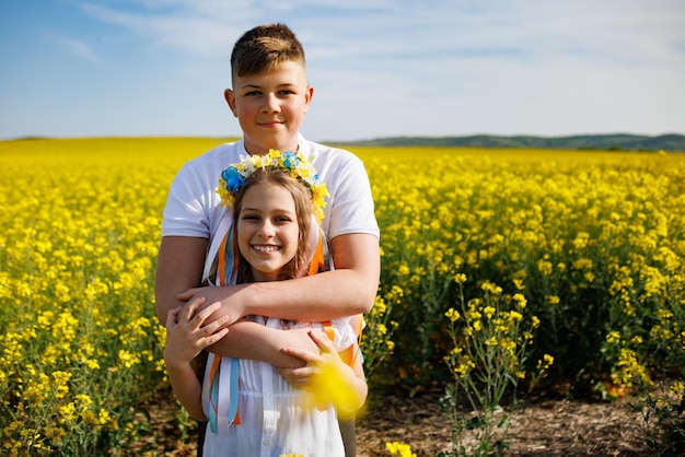 Photo brother hugs sister with ukrainian wreath and bouquet of flowers in hands in rapeseed field under sky