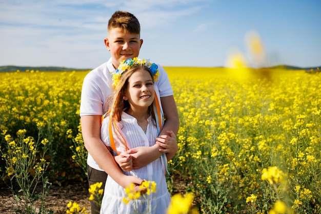 Brother hugs sister with Ukrainian wreath and bouquet of flowers in hands in rapeseed field under sky