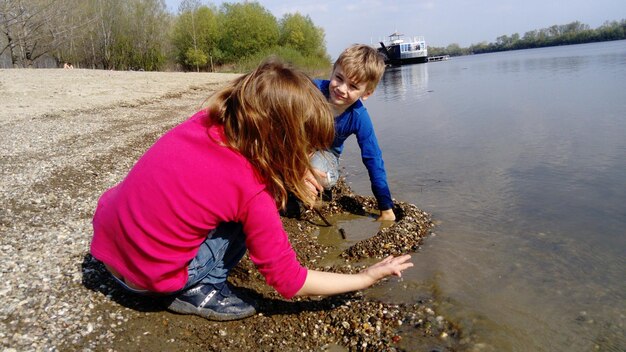 Brother and sister are playing by the river a boy and a girl\
are wearing jeans and longsleeved clothing early summer smiles and\
play by the water pink blouse and blue longsleeve danube river