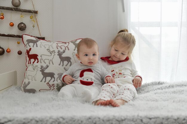 Брат и сестра в рождественской пижаме сидят на кровати с украшениями.