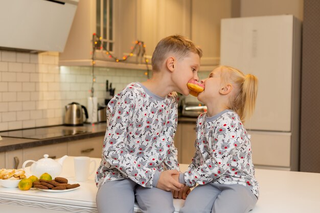 brother and sister in Christmas print clothes are biting one donut for two in a beautiful kitchen