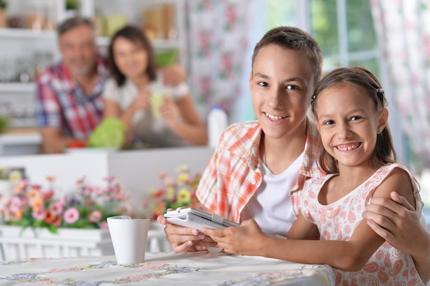 Brother and sister sitting at kitchen and using digital tablet with blurred parents on background