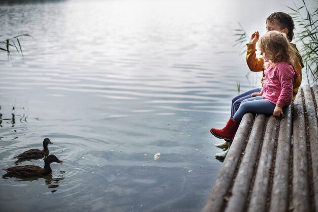 Photo brother and sister sitting on the river pier  and look at ducks in the water