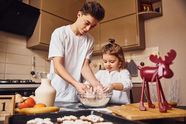 Brother with his little sister preparing food by using flour on kitchen and have fun.