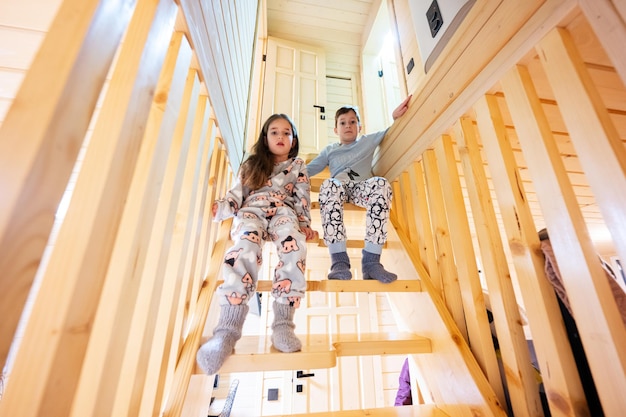 Brother with sister in pajama sit on stairs in cozy wooden tiny cabin house Life in countryside