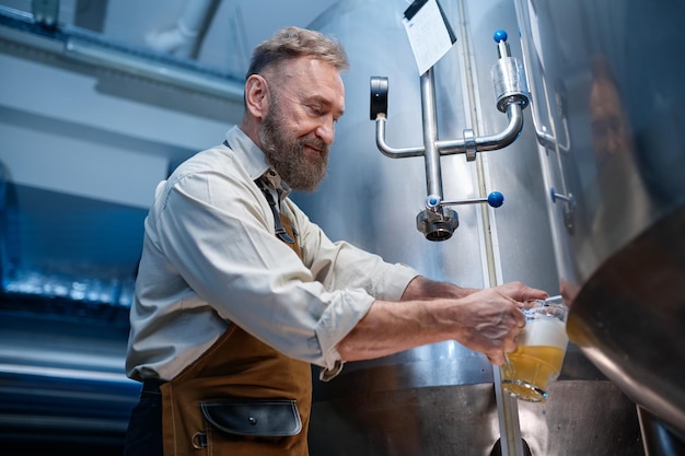 Brouwerij meester kwaliteitsproeverij van ambachtelijk bier in productiefaciliteit. Man brouwer vers gebrouwen bier of pils gieten in glas uit de metalen tank