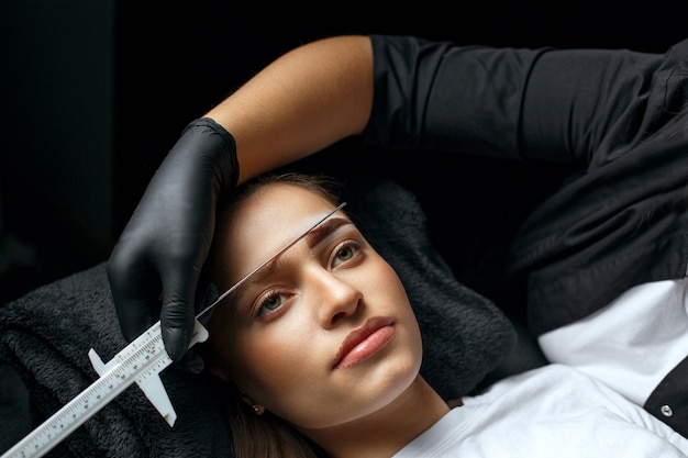 Photo brow stylist measuring eyebrows with the ruler before permanent makeup procedure in a beauty salon