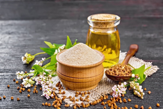 Brown buckwheat flour in a bowl, brown groats in a spoon, buckwheat flowers and leaves, oil in a glass jar on a burlap napkin on dark wooden board background