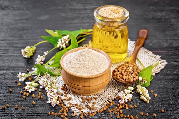 Brown buckwheat flour in a bowl, brown groats in a spoon, buckwheat flowers and leaves, oil in glass jar on a burlap napkin on wooden board background