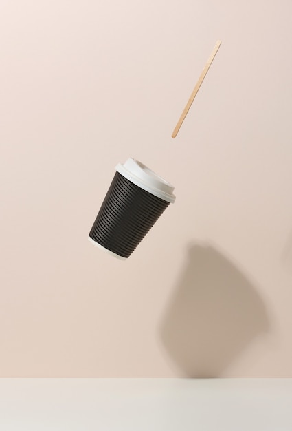 Photo brown cardboard cup with a white plastic lid and a wooden stirring stick levitate over a white table. beige background. coffee shop template