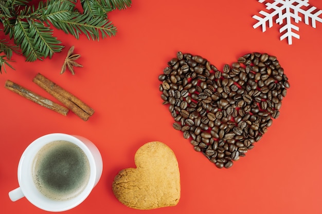 Brown coffee beans laid out in shape of heart with Christmas tree branches, cup of coffee, cookie on red background