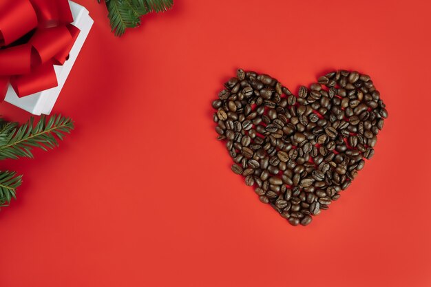 Brown coffee beans laid out in shape of heart with Christmas tree branches and gift box on red background, flat lay