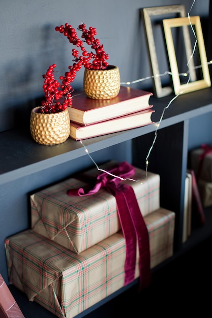 Фото Коричневые подарочные коробки с красной лентой, красная ягода илекса в золотых вазах на черной полке