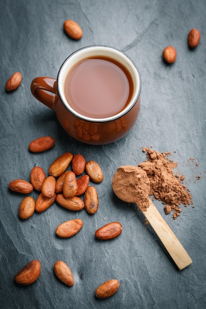 Brown cup of fresh cocoa drink with cocoa beans and powder in small wooden spoon on black stone background