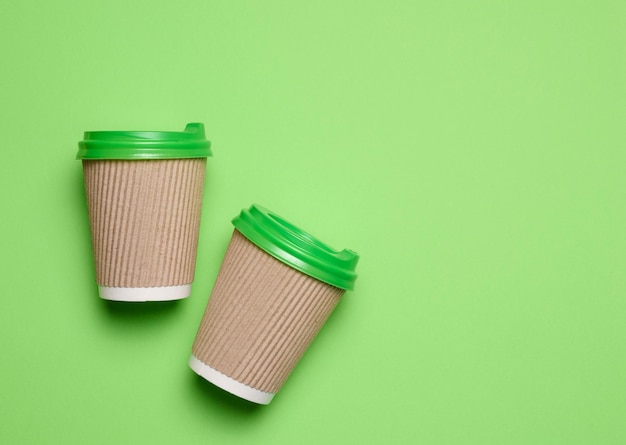 Brown disposable coffee and tea cups made of corrugated cardboard with green plastic lids on green background top view