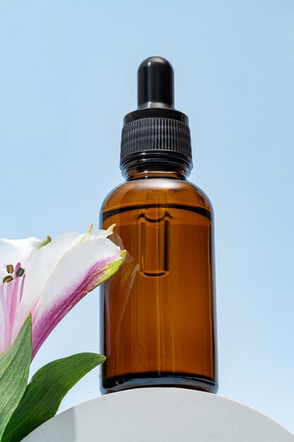 A brown dropper bottle of face oil displayed on a pedestal against a blue background with pink alstromeria flower cosmetic package unbranded mockup