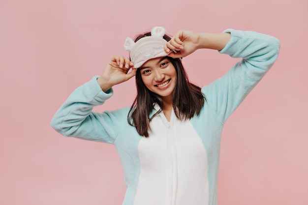 Brown-eyed brunette girl in cool mint soft pajamas puts on sleep mask and smiles sincerely on pink isolated wall