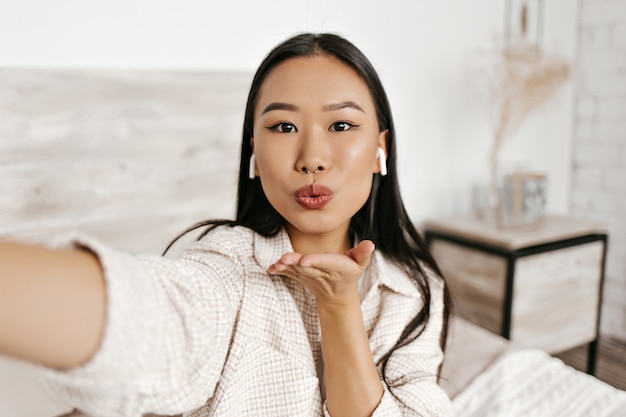 Brown-eyed brunette woman in plaid jacket takes selfie, blows kiss and listens to music in wireless headphones in cozy bedroom