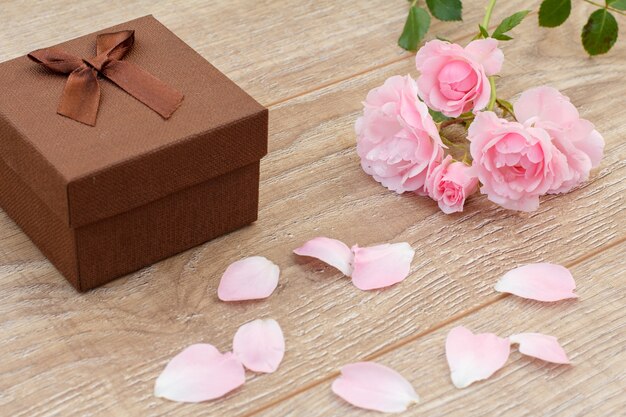 茶色のギフトボックス、バラの花びら、木製の背景に美しいピンクのバラ。休日に贈り物をするという概念。上面図。