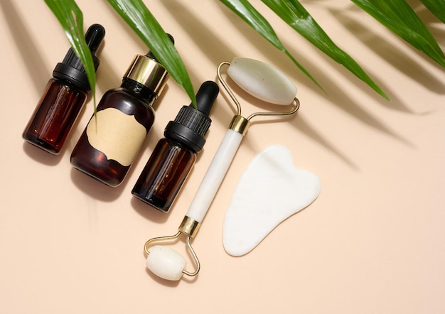 Brown glass bottle with a pipette a white stone hand massager and a scraper for cosmetic procedures on a beige background top view