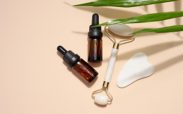 Brown glass bottle with a pipette a white stone hand massager and a scraper for cosmetic procedures on a beige background top view