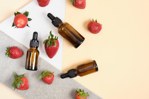 Brown glass bottles with pipette and strawberries on beige, white and grey background, top view