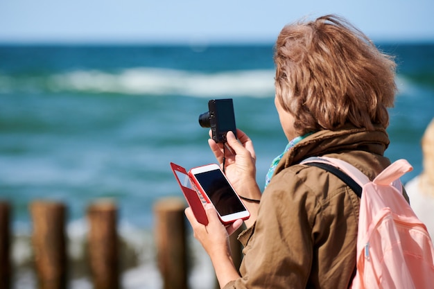 Brown-haired woman taking photos with digital camera, side view. Shooting video with digital camera, holding smartphone. Tourist photographing landscape by sea. Sightseeing, travel, tourism concept