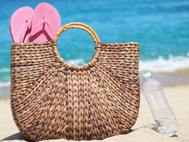 Brown natural wicker bag with flip flops on the beach next to a plastic water bottle