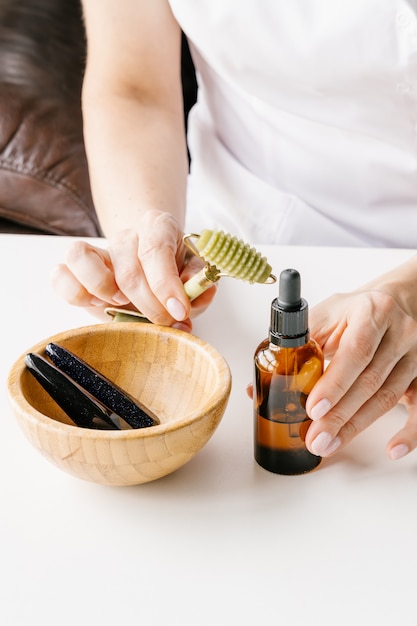 Brown plastic bottle for liquid cosmetics in woman's hands, green jade massage roller and wooden bowl with stone sticks on white table