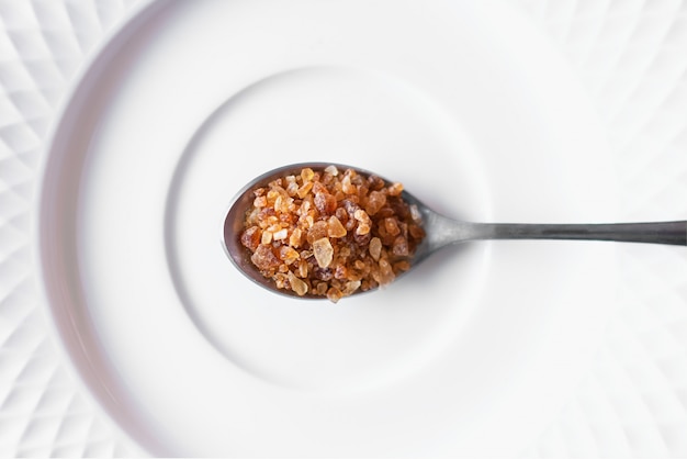 Brown rock sugar in spoon placed on white plate. Top view