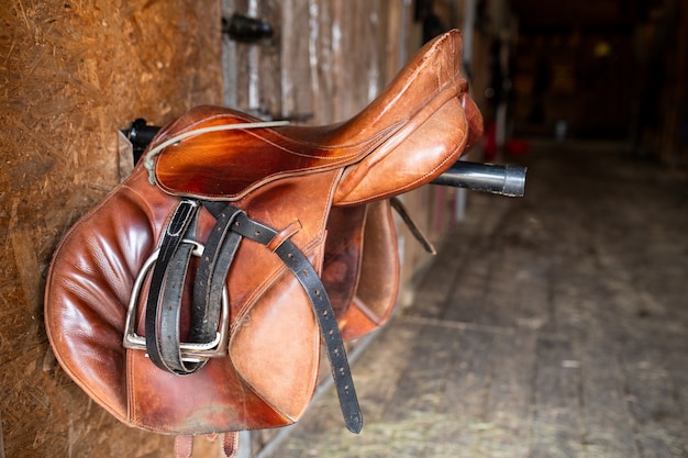 Photo brown shabby leather saddle with black bridles hanging on steel bar stuck out of wall inside stable