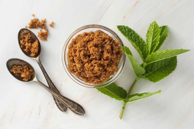 Brown sugar isolated in glass bowl, over styled marble background. Mint leaves. Top view, copy space.