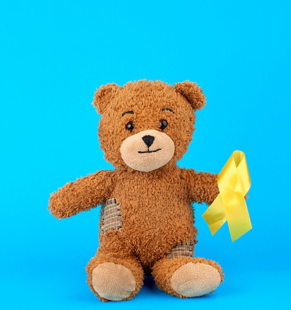 Brown teddy bear sits and holds in his paw a yellow silk ribbon on a blue background