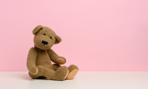 Brown teddy bear with patches sits on a white table, pink background, copy space