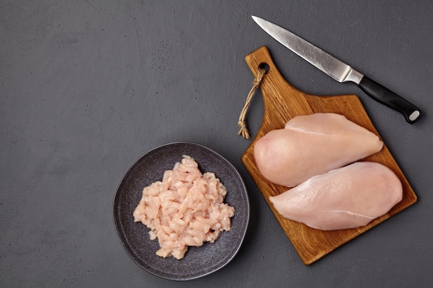 A brown wooden cutting board with pieces of chicken breast fillet and a bowl of chopped poultry meat and a knife on a dark concrete table Top view with copy space flat lay