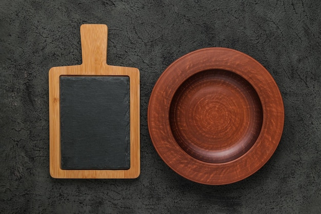 Brown wooden cutting board with slate stone empty brown clay plate on black stone table Top view with copy space Rustic dishes