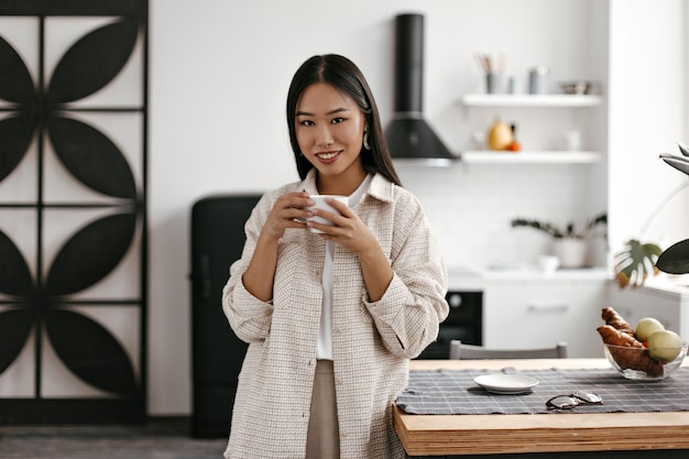 ベージュのカーディガンの茶色いブルネットの女性は居心地の良い部屋でお茶を飲む気分の良いアジアの女性は木製のテーブルに寄りかかって白いコーヒーカップを保持します