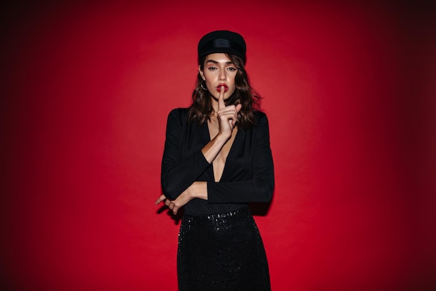 Browneyed woman in black cap and dress asks to be quiet Curly lady in stylish outfit poses on isolated red background