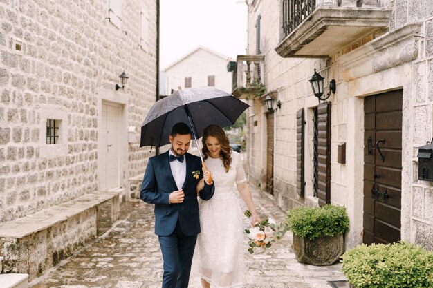 Bruid en bruidegom lopen over de kasseien van een oude straat onder een paraplu