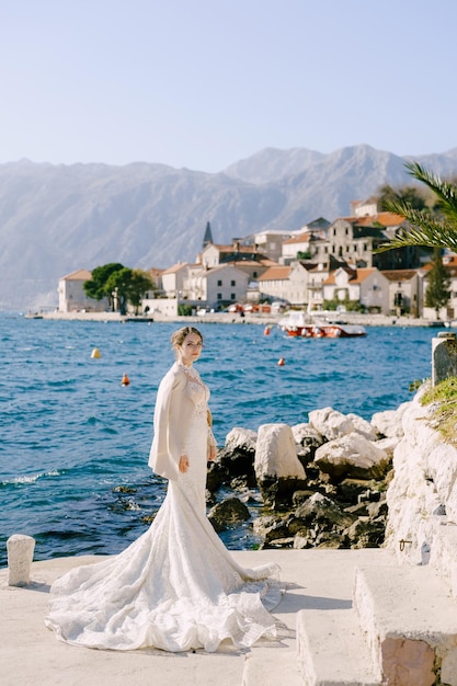 Bruid in een lange witte zeemeerminjurk staat op een pier voor de kust van perast