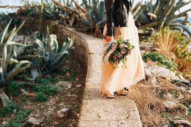 Bruid in een prachtige pastelkleurige jurk en een leren jas staat op een pad met een boeket bloemen