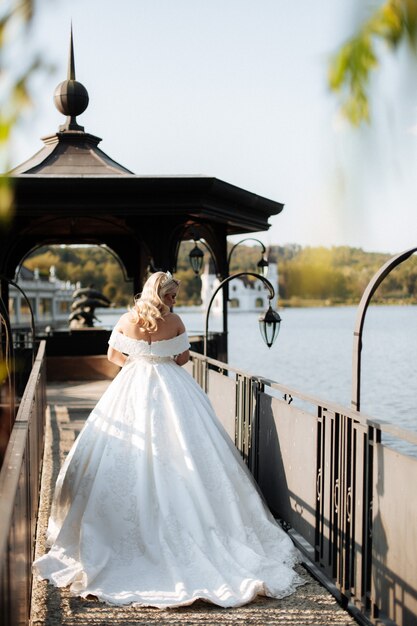 Foto bruid in een witte trouwjurk met een lange trein staat in een groen park, op de achtergrond zijn er bergen en een meer