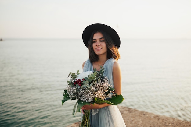 bruid in hoed met boeket bloemen aan de kust