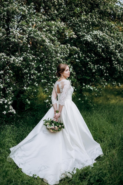 Bruid met een huwelijksboeket in het bos dichtbij de struiken die met witte bloemen bloeien