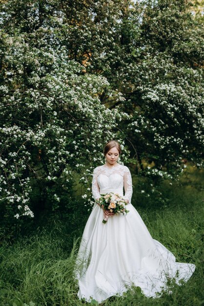 Bruid met een huwelijksboeket in het bos dichtbij de struiken die met witte bloemen bloeien