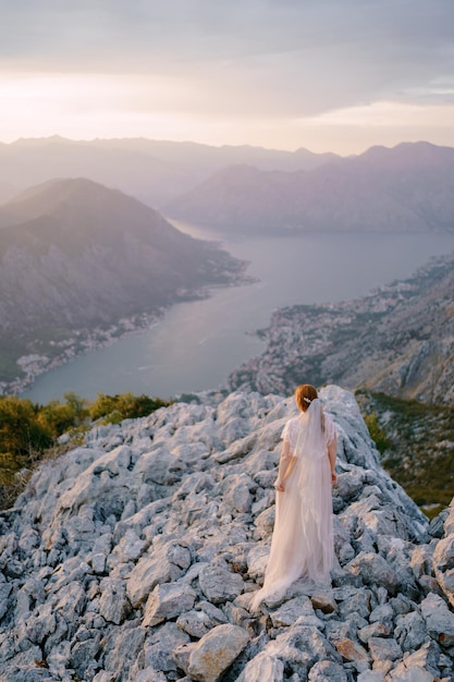 Bruid staat op de rotsen op de berg en kijkt neer op de baai