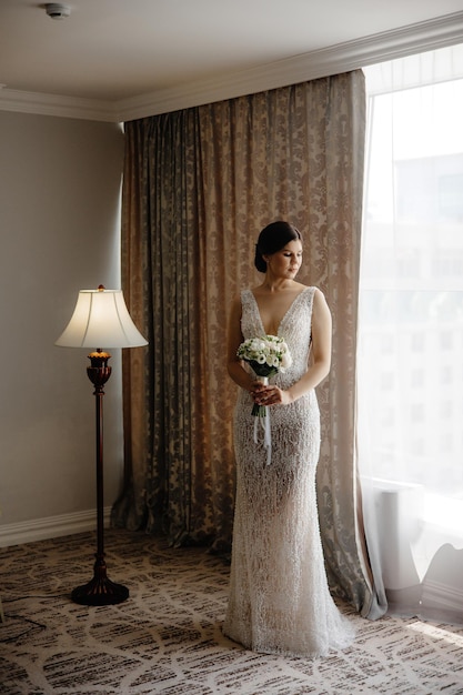 Bruid staat voor een raam met een witte trouwjurk