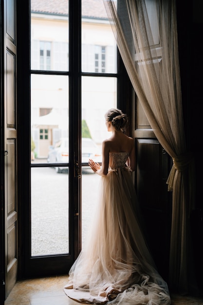 Bruid staat voor het grote raam van een oude villa en kijkt naar de binnenplaats van het Comomeer